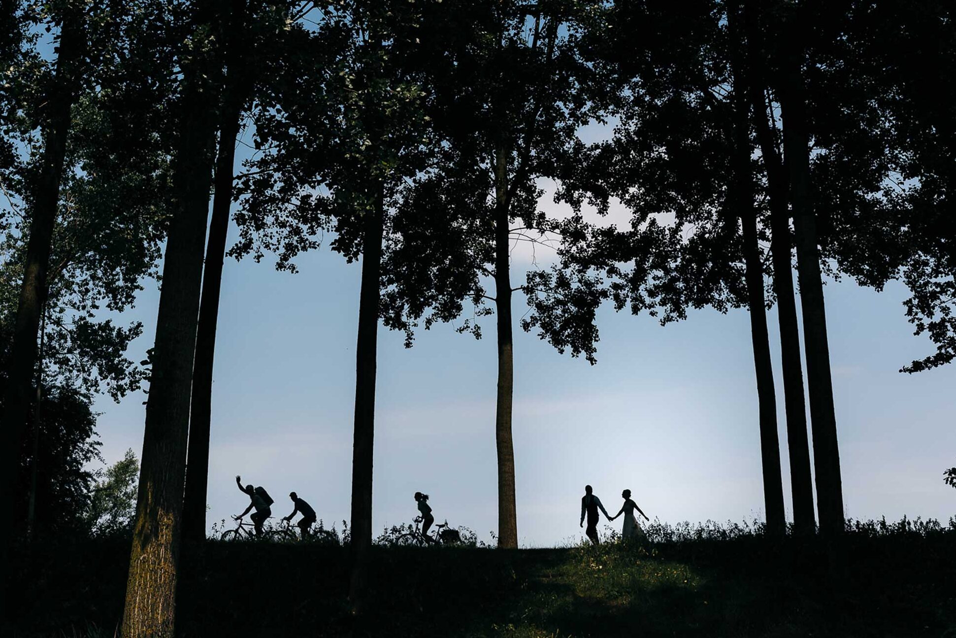 Een romantisch tafereel: het silhouet van een bruidspaar tussen de bomen, terwijl een voorbij fietsende fietser hen enthousiast begroet.