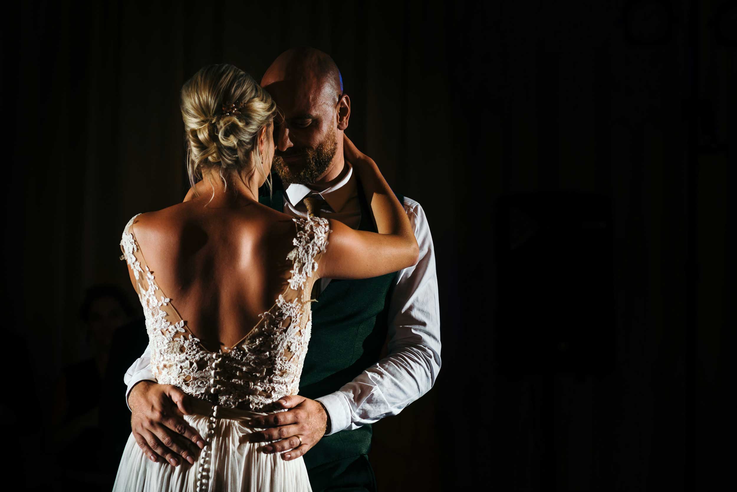 Een stralend bruidspaar betovert de dansvloer tijdens hun openingsdans. Het bruidspaar licht op tegen een sfeervolle, donkere achtergrond, waar het gebruik van flitslicht diepte en structuur toevoegt aan deze magische momentopname.