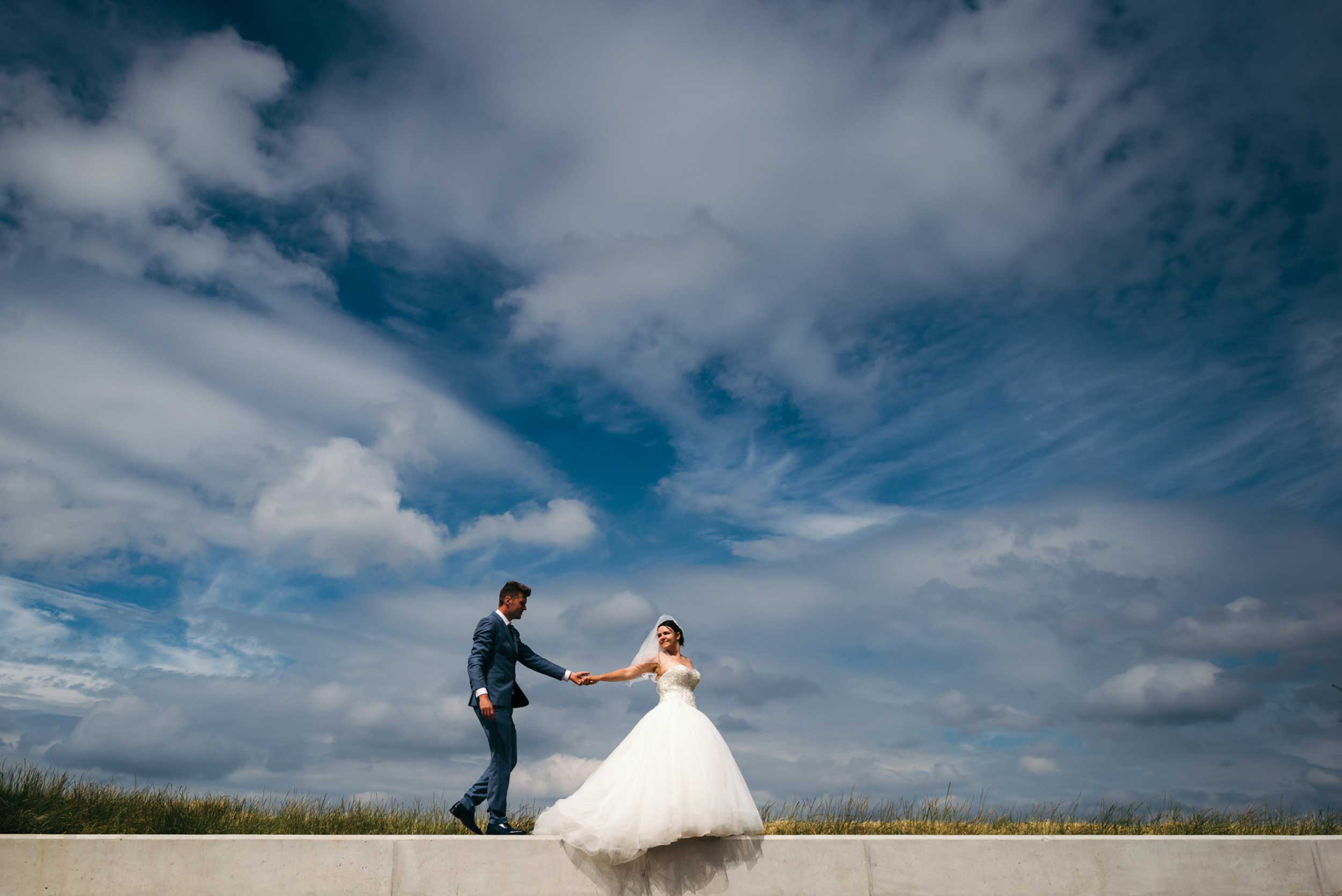 Een prachtig beeld van eenvoud: het bruidspaar wandelt hand in hand en steekt af tegen de zachte contouren van de wolken, een intiem moment van sereniteit en verbondenheid.
