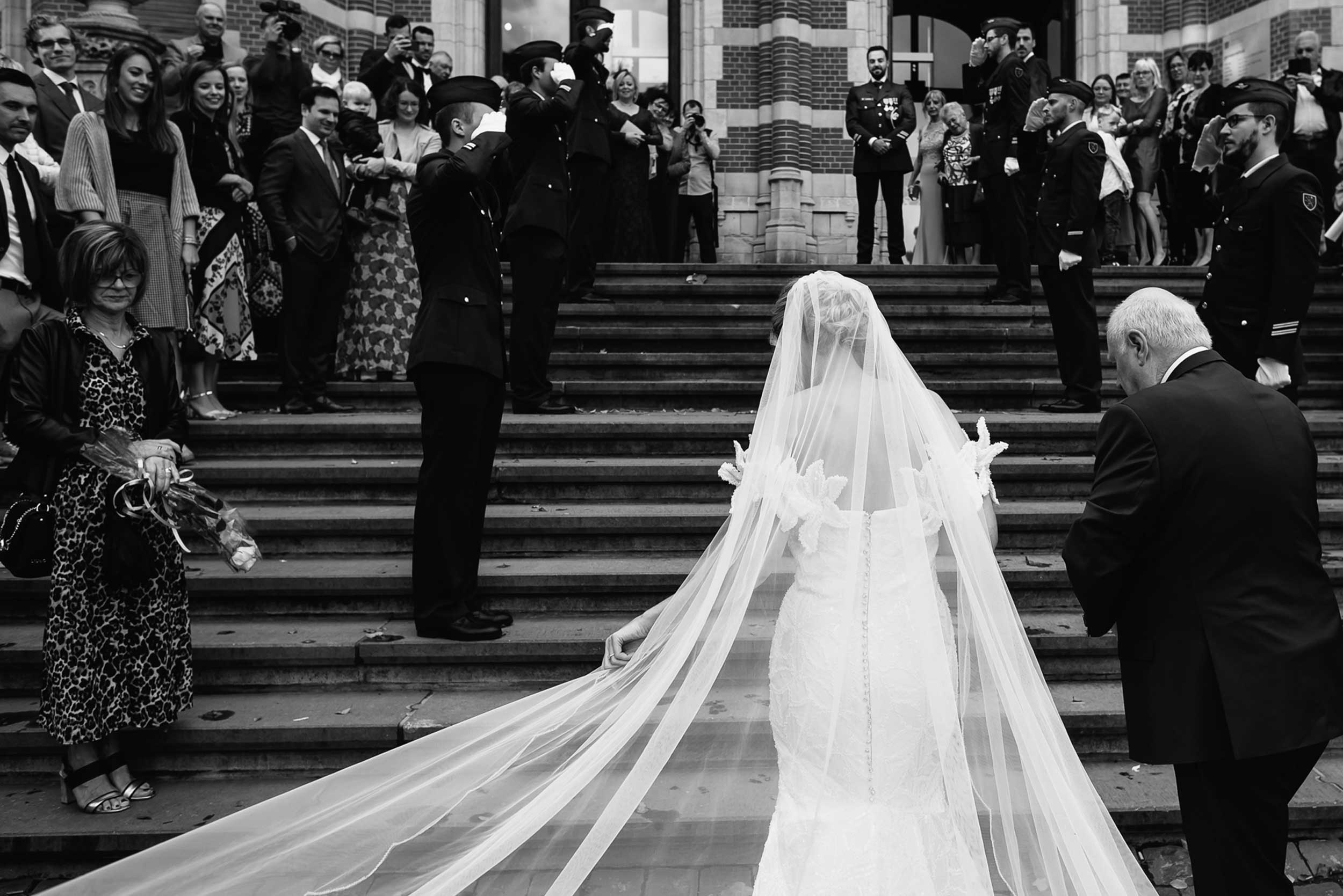 Eerbiedige Saluut: Militair huwelijk, het korps salueert terwijl de bruid de trap van het stadhuis opwandelt; sfeervol zwart-wit met de bruid in haar witte kleren die fel contrasteren met de donkere militaire uniformen.
