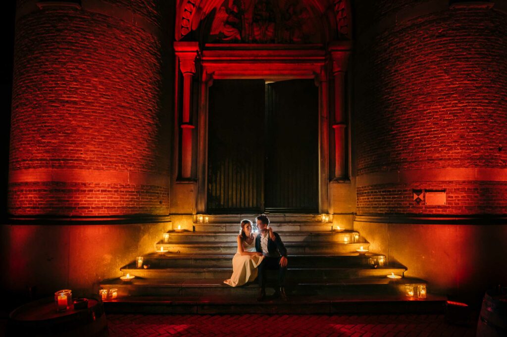 Sfeervolle nachtfoto: Bruidspaar zit op een verlichte trap voor een kerk, waarbij de kerk rood is uitgelicht en kaarsen de trap verlichten.