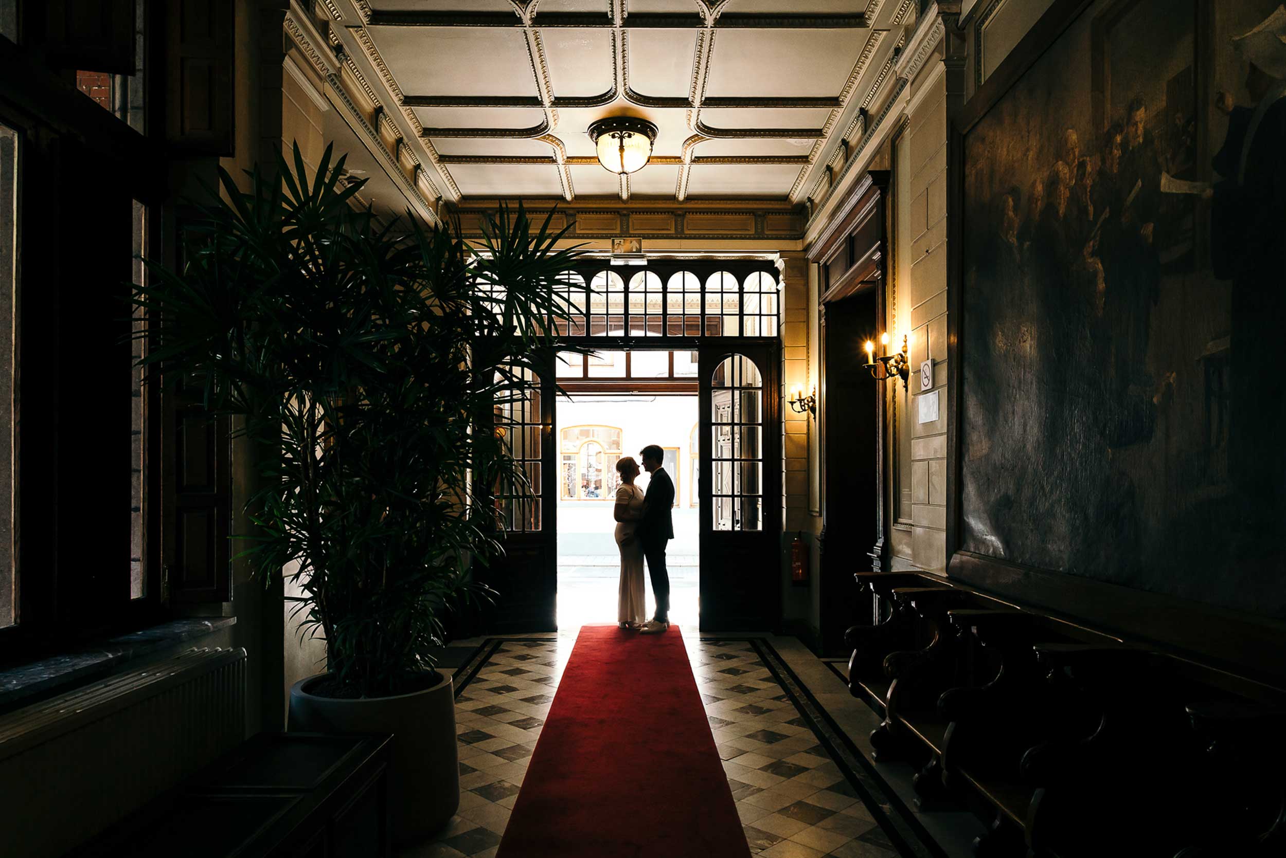 Prachtige silhouetfoto van bruidspaar, gefotografeerd van binnenuit naar buiten; strak retro interieur vormt een stijlvolle achtergrond.