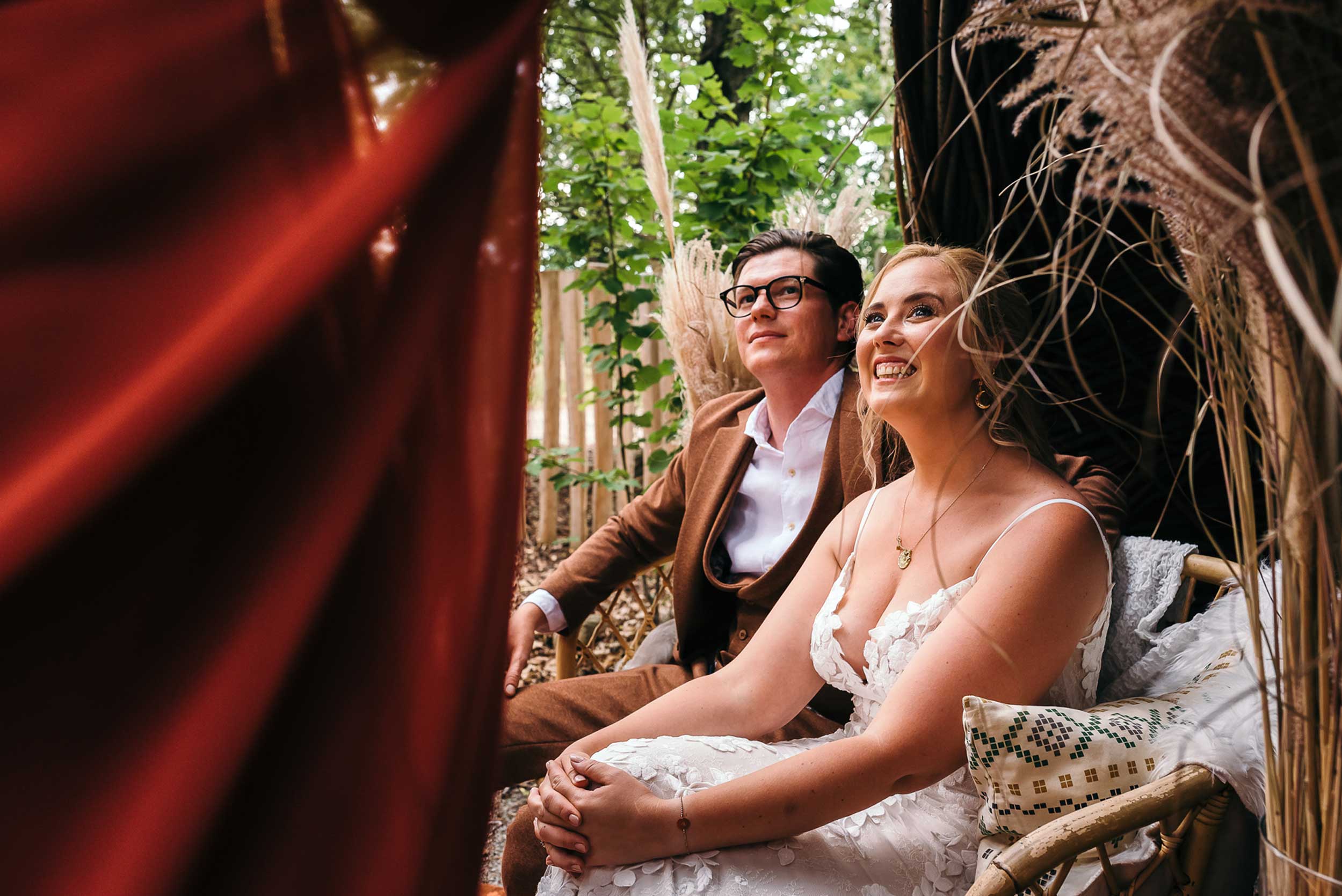 Bruid en bruidegom kijken vol verwachting naar de getuige in een visueel aantrekkelijke foto, waarbij het dieprode kleed van de getuige de helft van het beeld in beslag neemt.