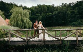 Prachtig bruidspaar staat elegant op een schilderachtig bruggetje over een rustige rivier in het idyllische Warredal.
